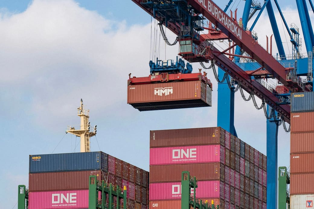 A shipping container suspended from a quay crane above a stacked container yard